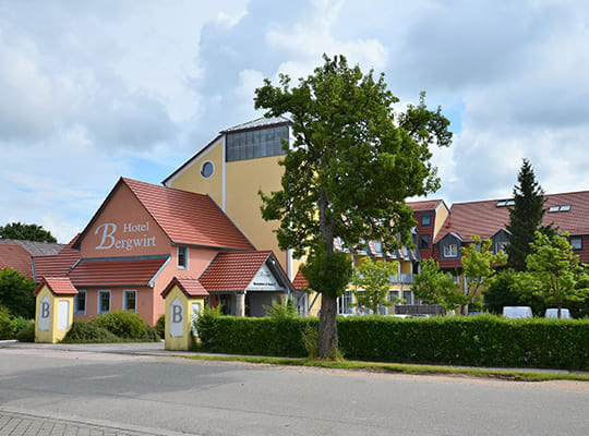 Außenansicht des Hotel-Gebäudes mit großem Baum und schöner Hecke davor.