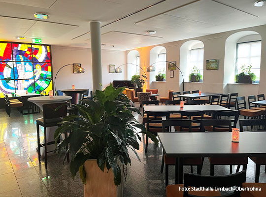 Länglicher Raum mit schwarzen Tischen, an welchen Stühle stehen. Am Ende des Raumes ist eine bunt gefärbte Glasscheibe