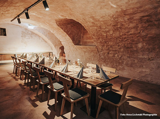 Gewölbekeller Optik mit langer Tischtafel im Innenraum, an dem Tisch stehen hölzerne Stühle